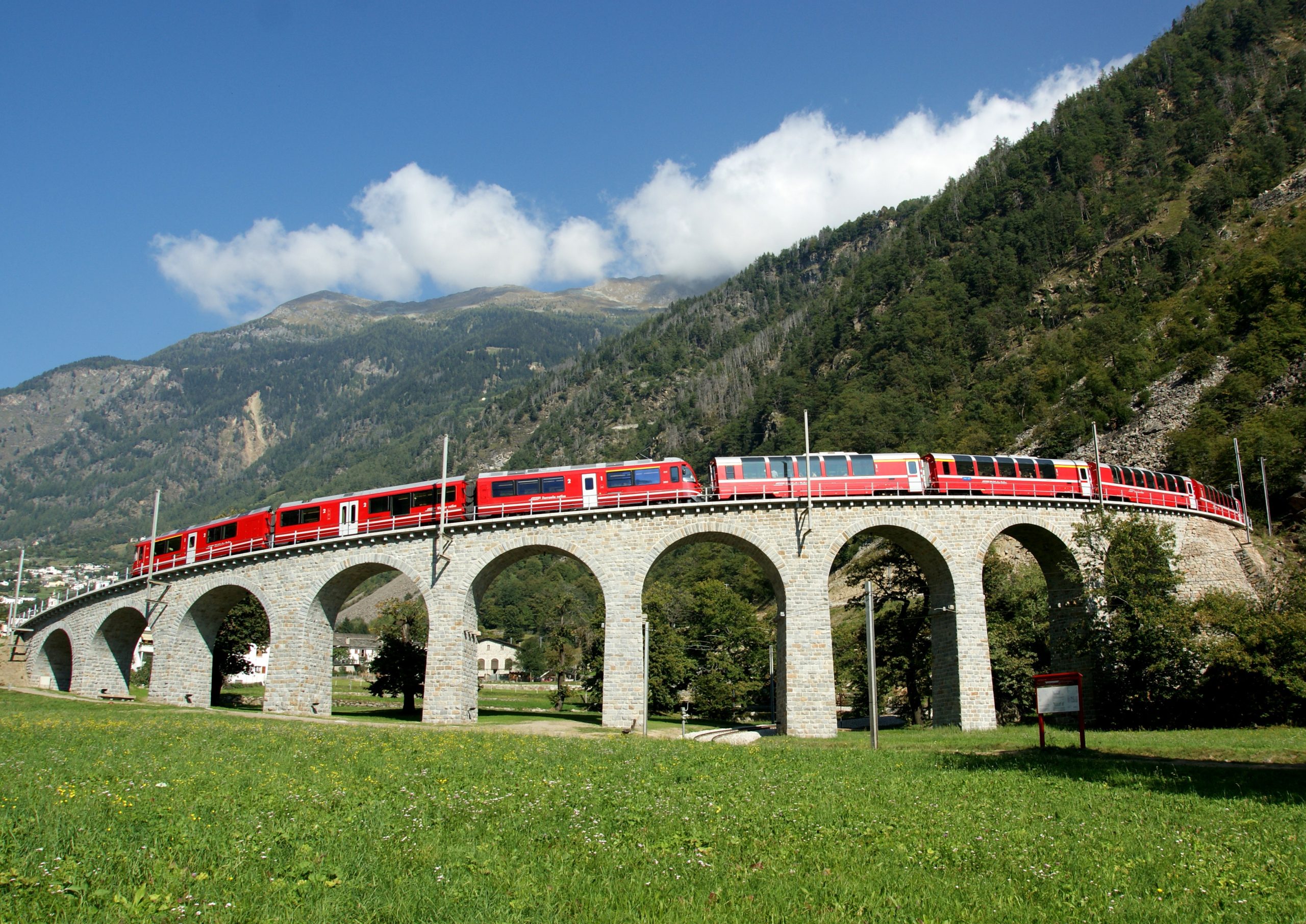 Treno rosso su viadotto in pietra tra prati e montagne alpine