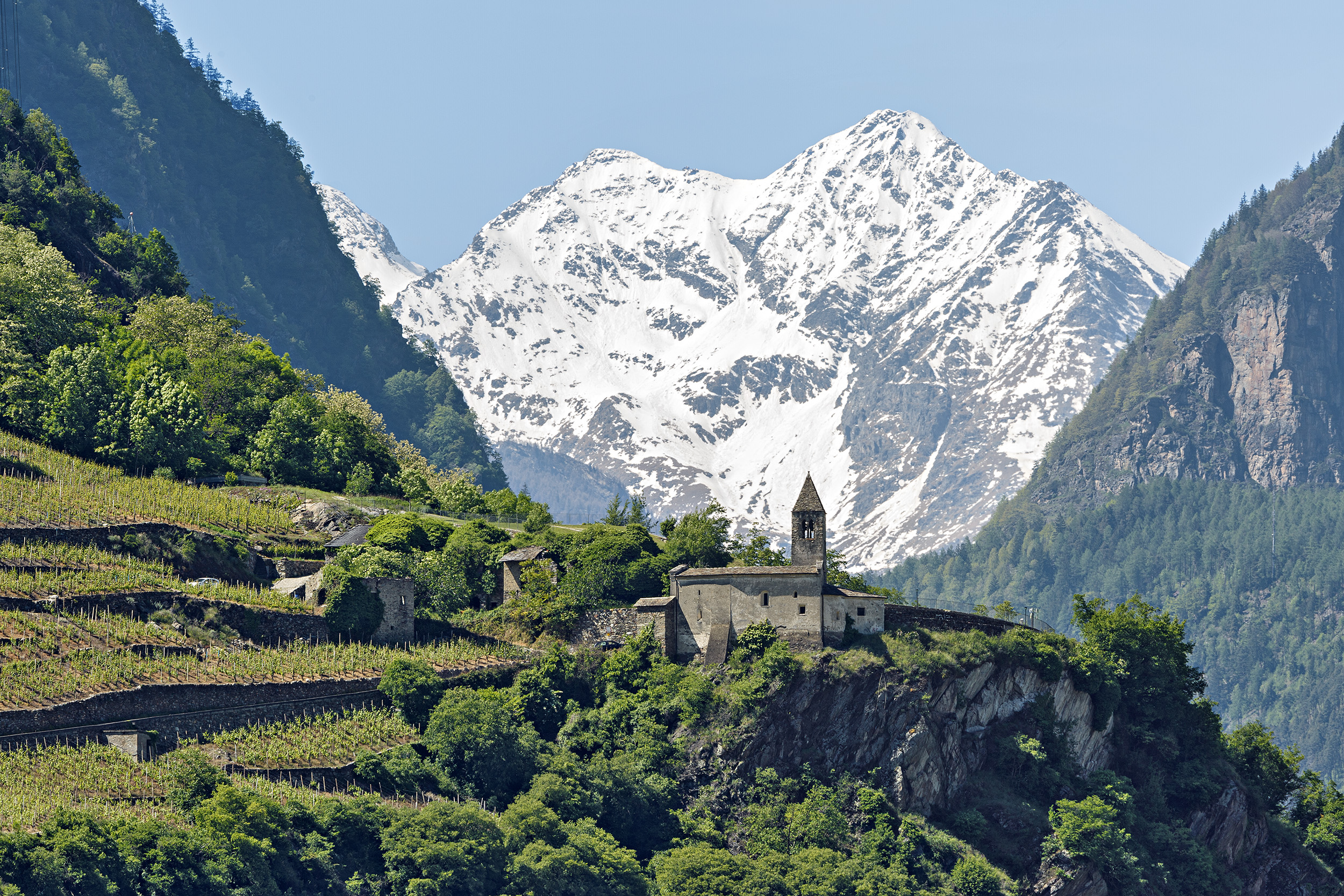 Église en pierre sur un promontoire entre des vignobles en terrasses avec des montagnes enneigées en arrière-plan