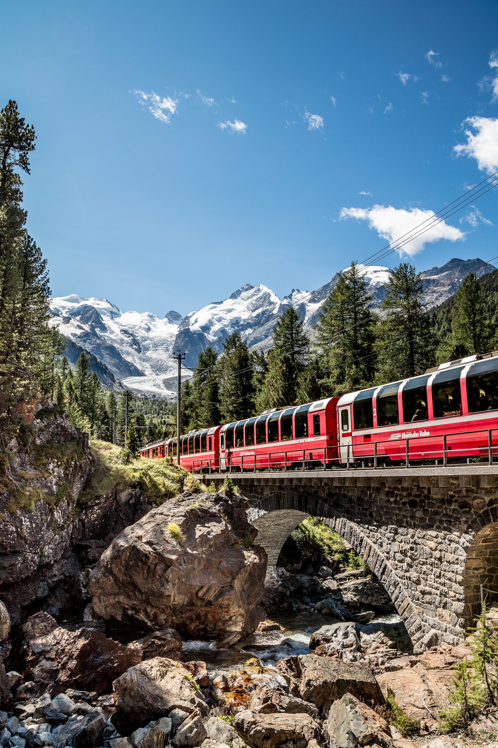 Treno rosso alpino su ponte in pietra tra boschi e montagne innevate