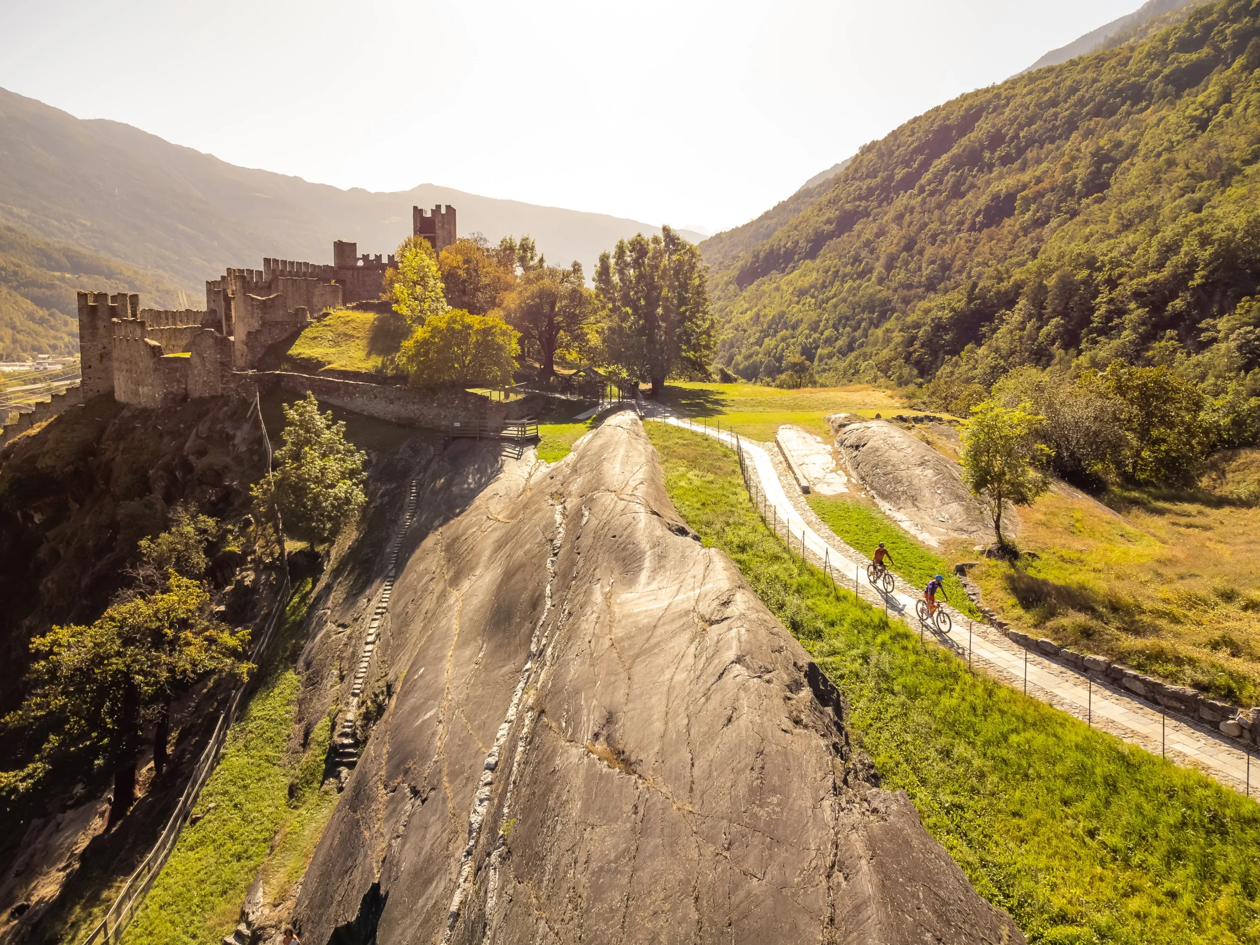 Burgruinen an einem felsigen Hang mit einem gewundenen Pfad und Radfahrern in einem grünen Tal unter bewaldeten Bergen.