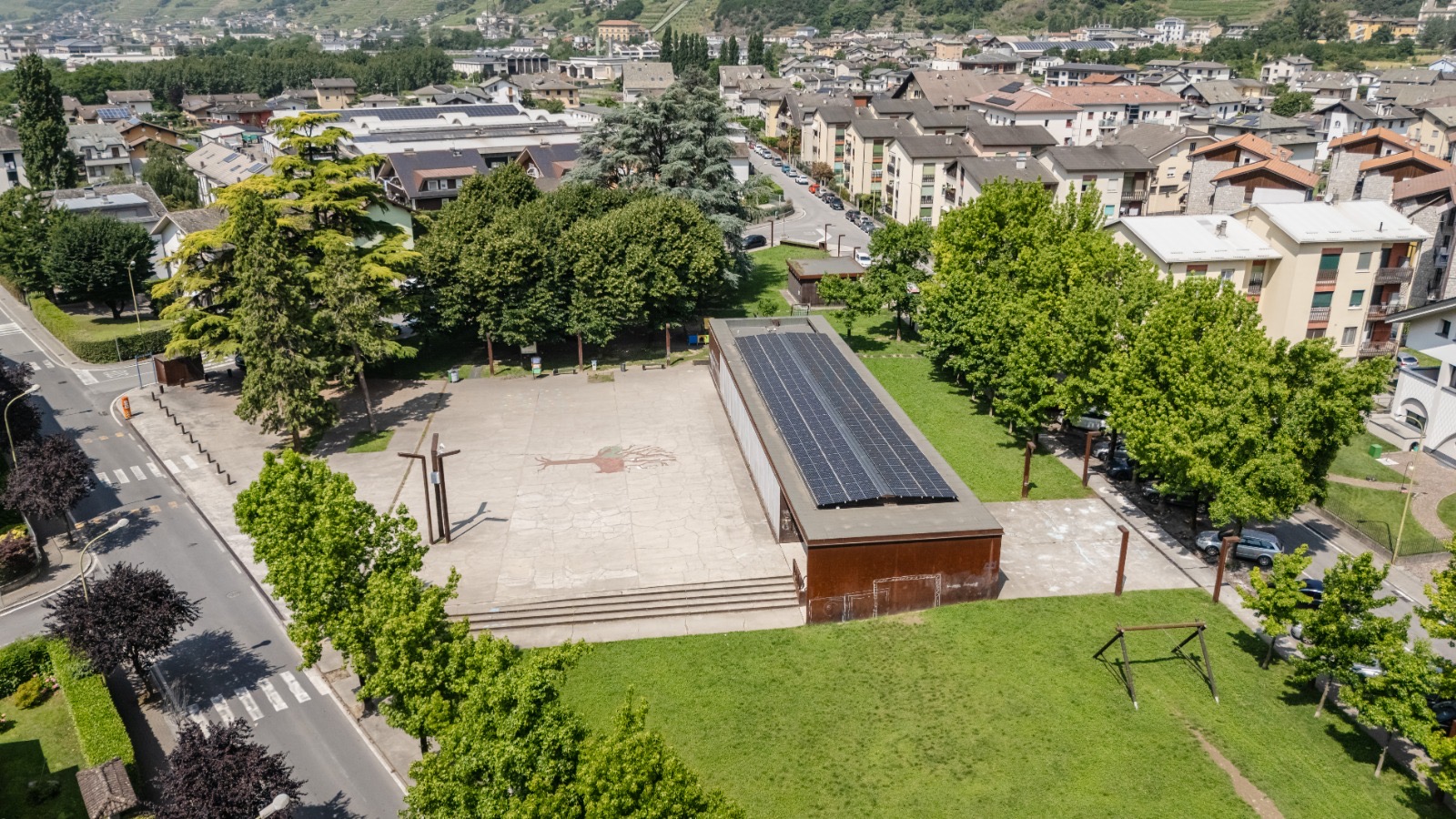 Aerial view of a town square with a long rectangular building covered in solar panels, surrounded by trees and residential buildings.