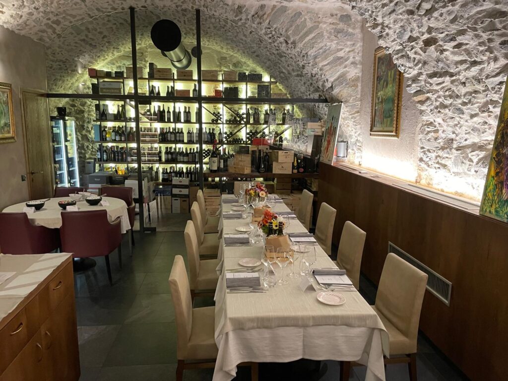 Cozy wine cellar restaurant with arched stone walls, illuminated wine shelves, and a long white-tablecloth dining table set for dinner.